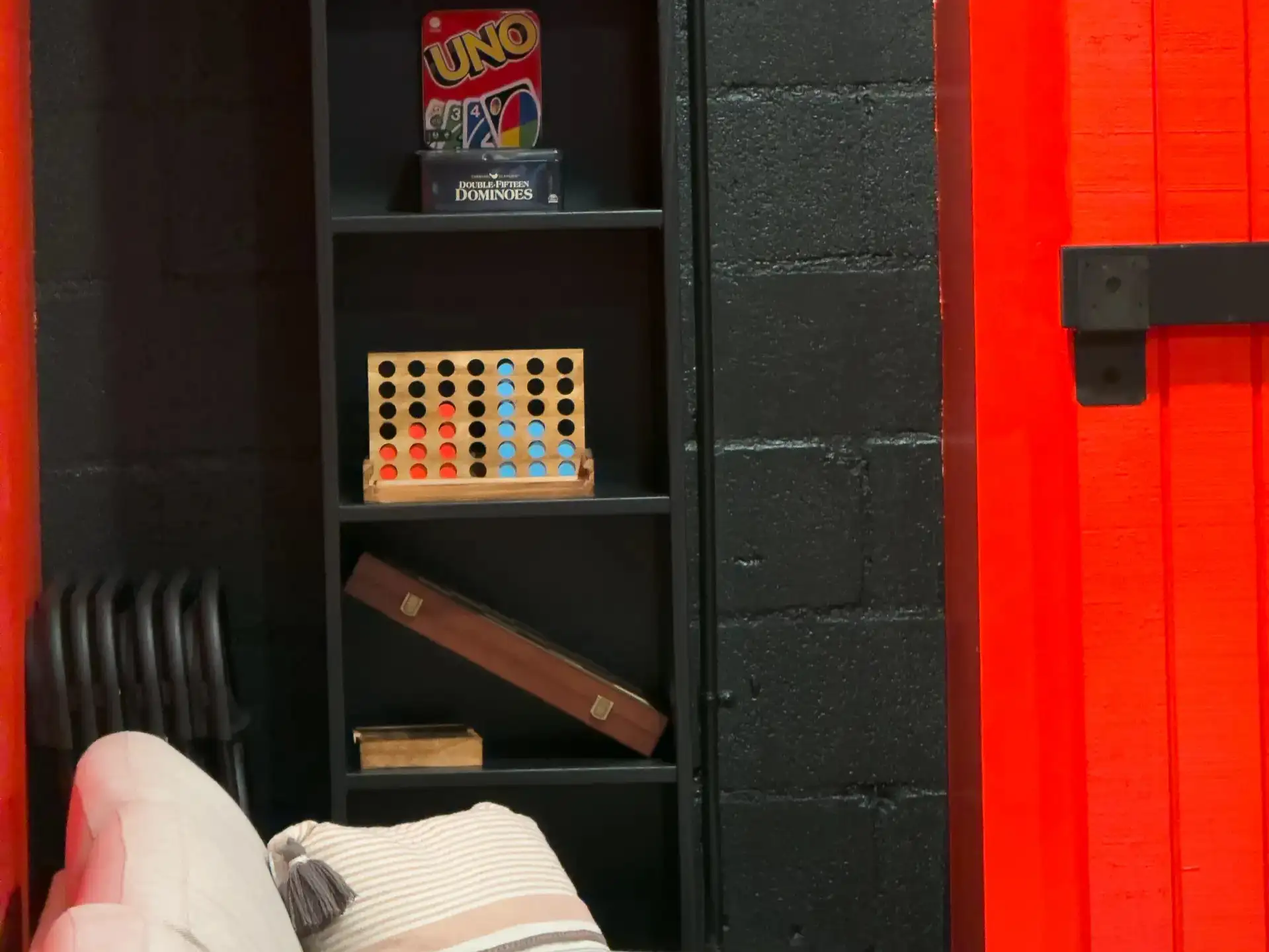 A shelf of board games and puzzles beside the party room's red plank wall, with the lounge sofa in front